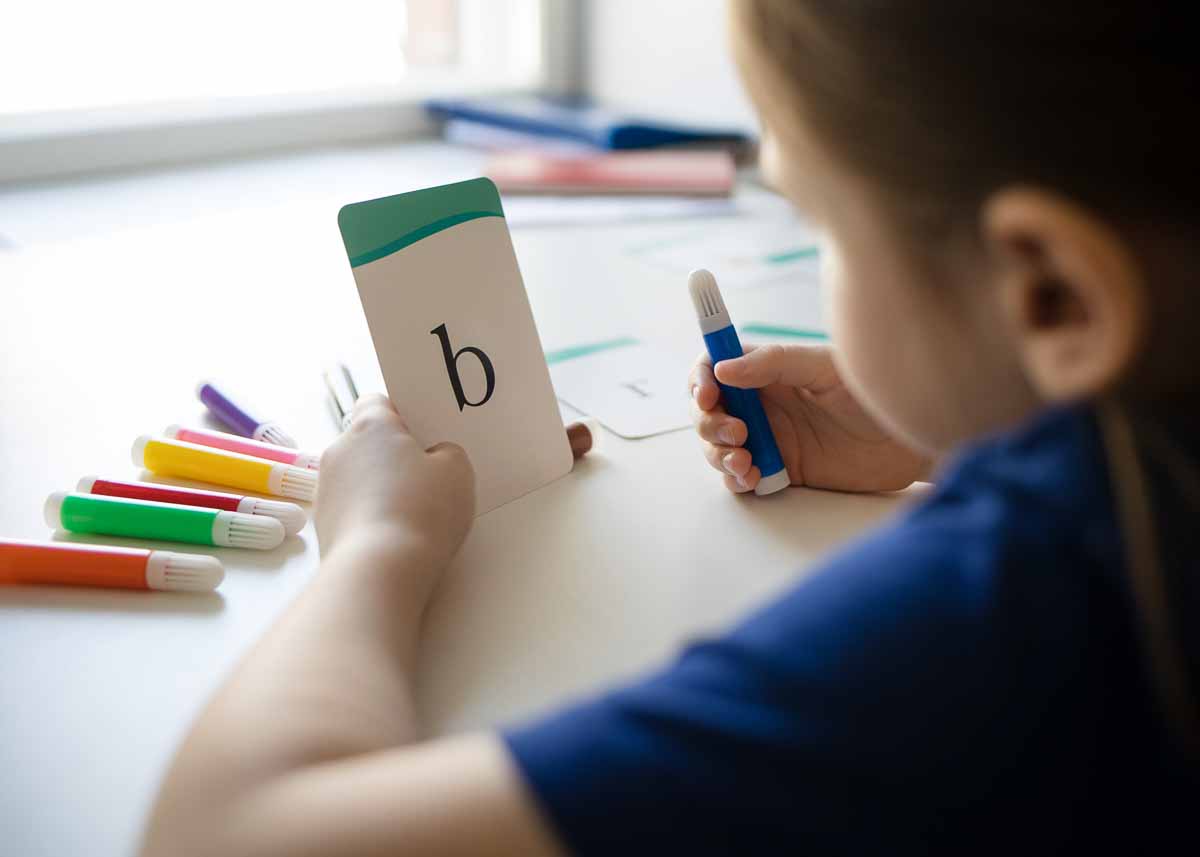 aula de alfabetização, menina segura um cartão branco com borda verde e a letra b escrita em uma mão e segura caneta hidrográfica com a outra.