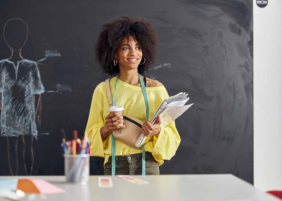 Sorridente jovem professora negra com fita métrica, xícara e papéis fica à mesa contra o quadro-negro com figura humana desenhada em vestido na aula de costura