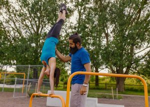 Mulher se equilibra em uma barra de ferro com as mãos enquanto é auxiliada por homem, exemplo de calistenia, nicho do mercado de trabalho para educação física