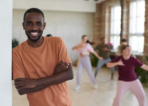 Homem sorri para câmera enquanto grupo de mulheres dançam ao fundo, exemplo de dança, nicho do mercado de trabalho para educação física