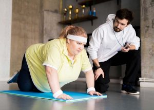Homem observa mulher obesa fazer flexões, exemplo de atividade de emagrecimento, nicho do mercado de trabalho para educação física