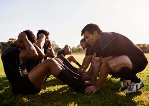 Atletas ajudando uns aos outro a fazer abdominais, exemplo de preparação física para esportes, nicho do mercado de trabalho para educação física