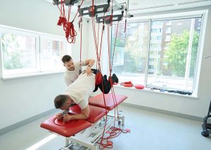Homem fazendo exercício de descompressão de coluna monitorado por outro homem, exemplo de atividade de reabilitação, nicho do mercado de trabalho para educação física